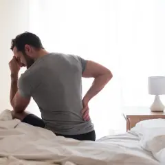 Homme assis sur un lit, main sur le front et l'autre sur le bas du dos, cherchant comment dormir avec une sciatique.