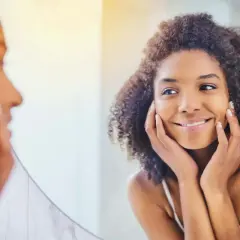 Une femme aux cheveux bouclés se regarde dans un miroir, les mains posées sur ses joues, comme pour se masser le visage. Elle sourit doucement.