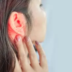 Une femme se touche l'oreille, une zone rouge vif indiquant une douleur ou une inflammation. Un point noir est visible dans l'oreille.