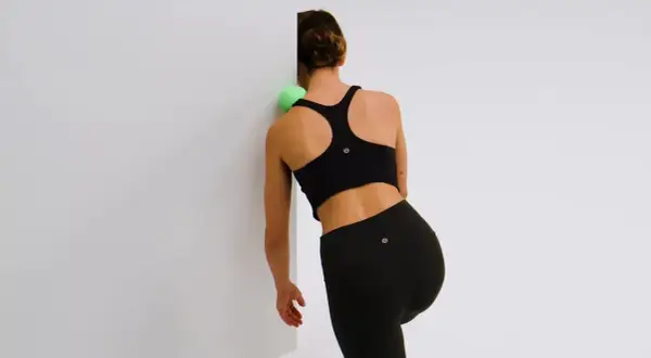 Femme utilisant une balle verte pour masser et muscler ses omoplates contre un mur blanc.