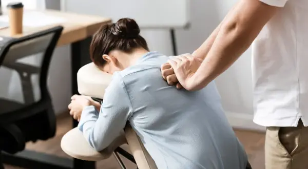 Un masseur offre des massages en entreprise à une femme assise sur une chaise ergonomique, soulageant les tensions du dos.