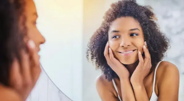 Une femme aux cheveux bouclés se regarde dans un miroir, les mains posées sur ses joues, comme pour se masser le visage. Elle sourit doucement.