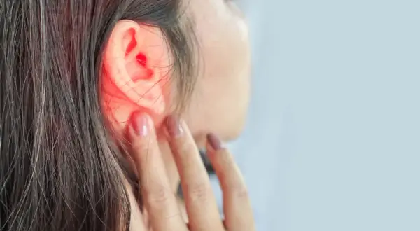 Une femme se touche l'oreille, une zone rouge vif indiquant une douleur ou une inflammation. Un point noir est visible dans l'oreille.