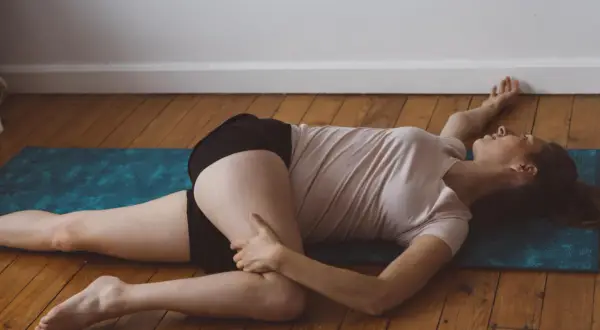 Une femme s'étire le dos à deux sur un tapis de yoga bleu. Elle est allongée sur le côté, une jambe pliée et l'autre tendue.