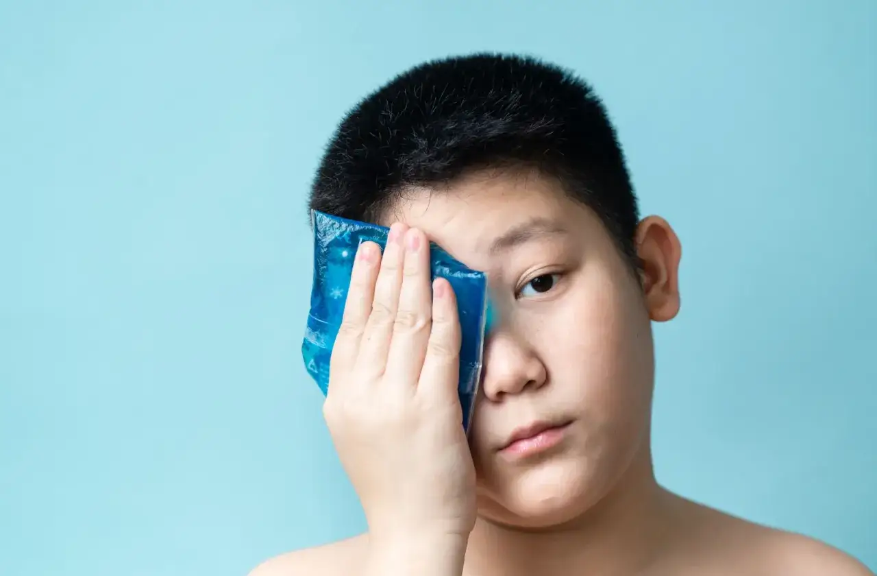 Un enfant applique une compresse froide sur son &oelig;il pour soulager un visage gonfl&eacute;, un rem&egrave;de de grand-m&egrave;re efficace.