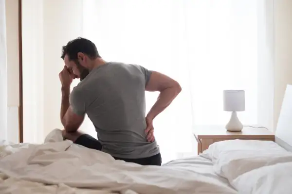 Homme assis sur un lit, main sur le front et l'autre sur le bas du dos, cherchant comment dormir avec une sciatique.