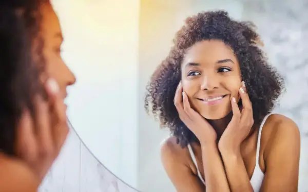 Une femme aux cheveux bouclés se regarde dans un miroir, les mains posées sur ses joues, comme pour se masser le visage. Elle sourit doucement.